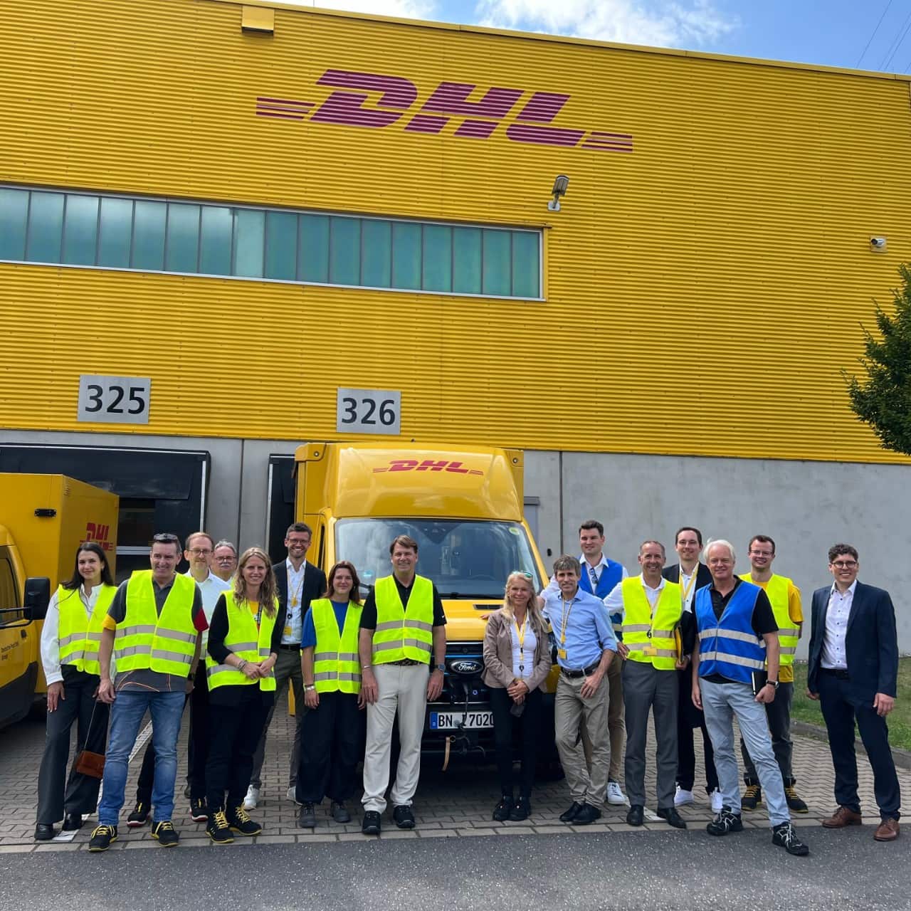 A group of people, most of them in neon yellow vests, pose in front of the DHL building.